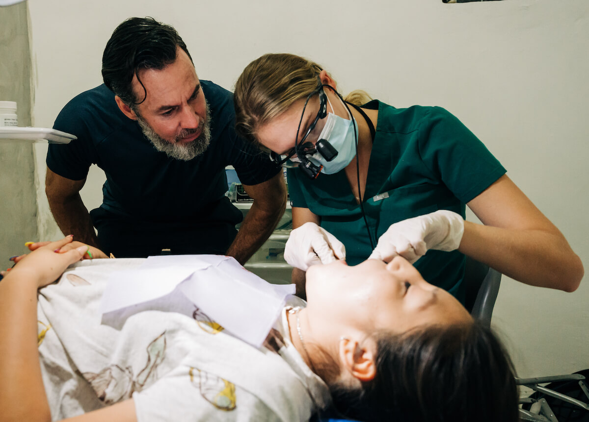 A mentor dentist oversees his mentee as she performs a dental checkup on a pediatric patient on a Humanitarian Trip. Learning on the job may be better for some new dentists vs AEGD vs GPR.